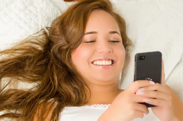Jovem morena na cama, descansando confortavelmente em lençóis brancos e travesseiro, segurando o telefone celular sorrindo — Fotografia de Stock