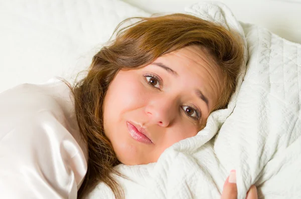 Jonge brunette vrouw headshot in bed liggen met het hoofd rust op een kussen, gefrustreerd ziek gelaatsuitdrukking — Stockfoto