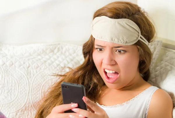 Mujer morena joven en la cama, descansando cómodamente sobre sábanas blancas y almohada, sosteniendo el teléfono móvil, expresión facial sorprendida —  Fotos de Stock