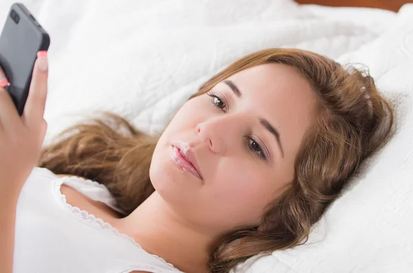 Mujer morena joven en la cama, descansando cómodamente sobre sábanas blancas y almohada, sosteniendo el teléfono móvil sonriendo — Foto de Stock