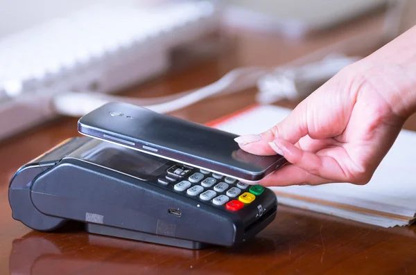 Uma mão de mulher segurando um telefone inteligente perto de uma máquina de cartão de crédito em uma mesa de madeira — Fotografia de Stock