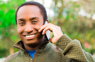 Yakışıklı İspanyol siyah adam açık park alanında yeşil kazak giyen kulak telefonu tutarak ve gülerken mutlu bir şekilde konuşuyor