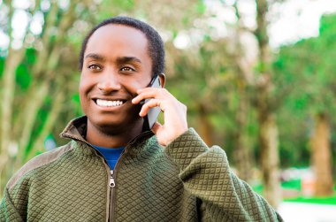 Yakışıklı İspanyol siyah adam açık park alanında yeşil kazak giyen kulak telefonu tutarak ve gülerken mutlu bir şekilde konuşuyor