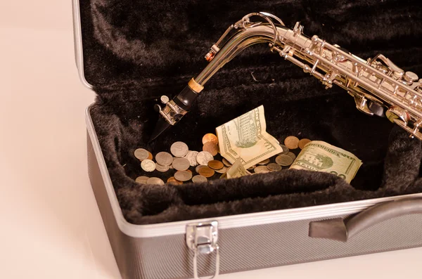 Glänsande saxofon liggande över öppna instrumentala hölje med svart sammet interiör och hög med pengar inuti — Stockfoto