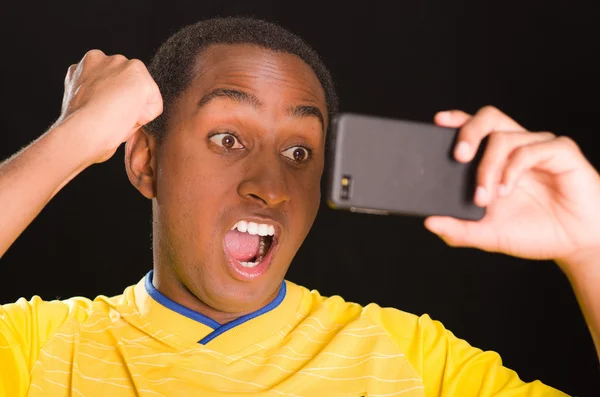 Headshot dark skinned male wearing yellow football shirt in front of black background, holding up mobile phone watching screen and cheering enthusiastically — Stock Photo, Image