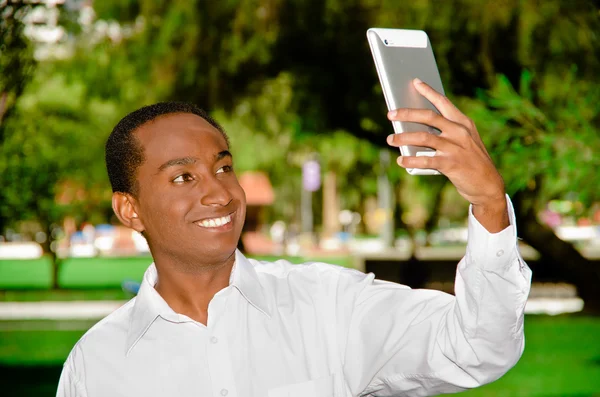 Bello ispanico nero uomo che indossa camicia bianca in zona parco all'aperto tenendo in mano tablet e guardando lo schermo come nel prendere selfie — Foto Stock