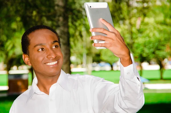 Bello ispanico nero uomo che indossa camicia bianca in zona parco all'aperto tenendo in mano tablet e guardando lo schermo come nel prendere selfie — Foto Stock