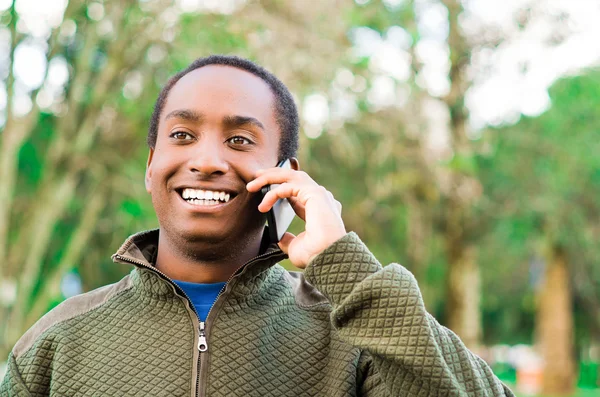 Hombre negro hispano guapo usando suéter verde en el área del parque al aire libre sosteniendo el teléfono al oído y hablando felizmente mientras se ríe — Foto de Stock