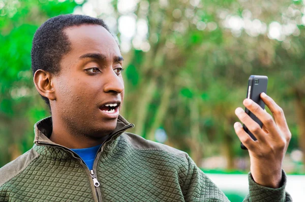 Handsome hispanic black man wearing green sweater in outdoors park area holding up phone and watching screen as in taking selfie — Stock Photo, Image