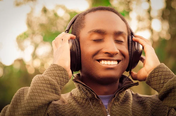 Hombre negro hispano guapo usando suéter verde en el área del parque al aire libre, manos sosteniendo auriculares cubriendo oídos y sonriendo disfrutando un poco de música — Foto de Stock