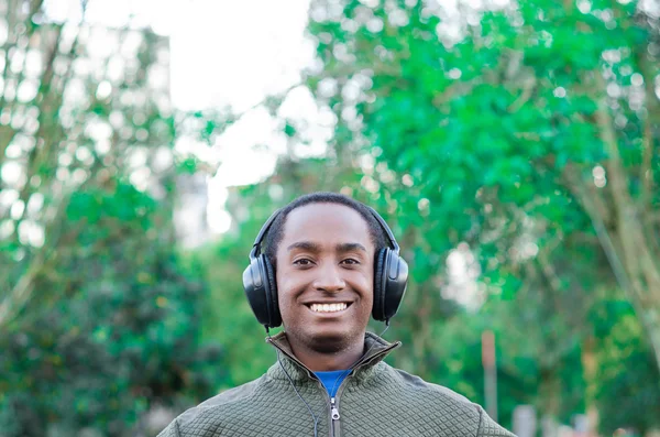Hombre negro hispano guapo con suéter verde en la zona del parque al aire libre, auriculares en las orejas que cubren y sonriendo disfrutando de un poco de música — Foto de Stock
