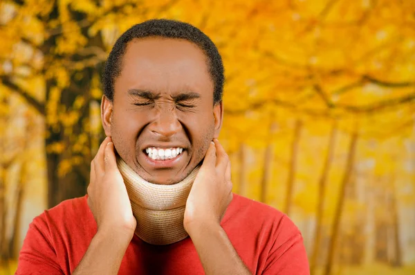 Injured young positive black hispanic male wearing neck brace, holding hands in pain around support making faces of agony, yellow abstract background — Stock Photo, Image