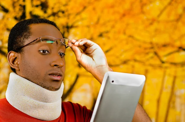 Jovem ferido positivo hispânico preto masculino vestindo cinta de pescoço, segurando tablet e leitura de tela levantando óculos na testa, fundo abstrato amarelo — Fotografia de Stock