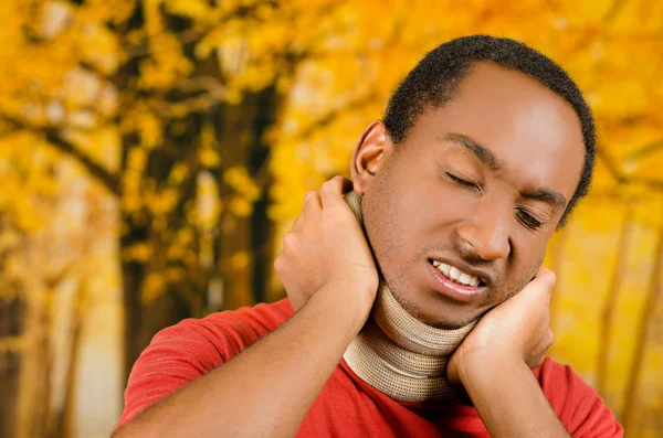 Injured black hispanic male wearing neck brace, holding hands in pain around support making faces of agony, yellow abstract background