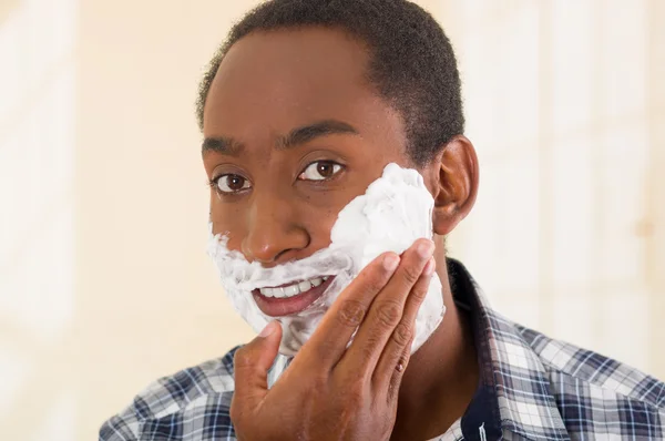 Jeune homme portant blanc bleu chemise à motif carré appliquer de la mousse à raser sur le visage en utilisant les mains, en regardant dans la caméra — Photo