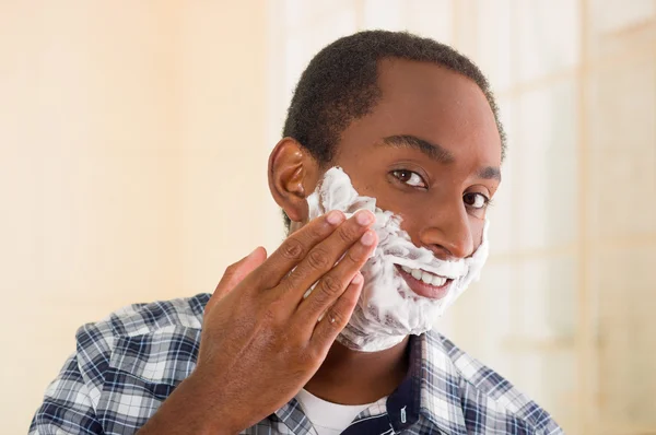 Молодой человек в белой синей квадратной рубашке наносит пену для бритья на лицо руками, смотрит в камеру — стоковое фото
