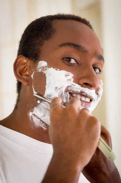 年轻男子穿着白色T恤，用围巾刮胡子，脸上涂满泡沫，看着相机 — 图库照片
