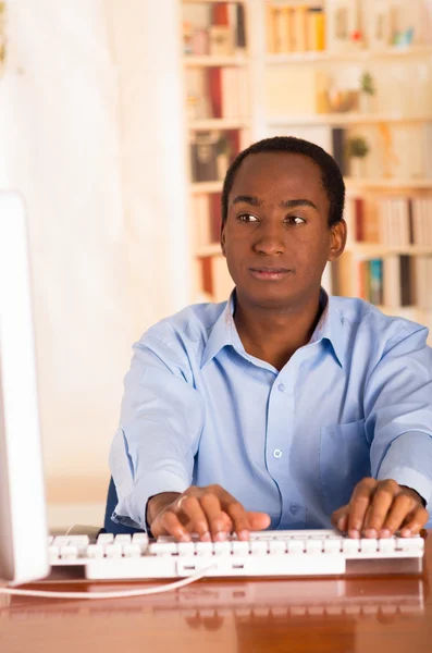 Mladý pohledný muž v modré kancelářské košili sedí u počítačové psací stůl a vypadá nenápadně — Stock fotografie