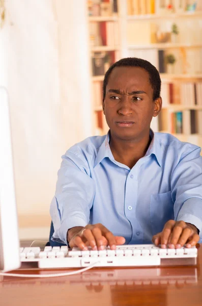 Joven hombre guapo con camisa de oficina azul sentado junto al escritorio de la computadora escribiendo y mirando sin inspiración —  Fotos de Stock