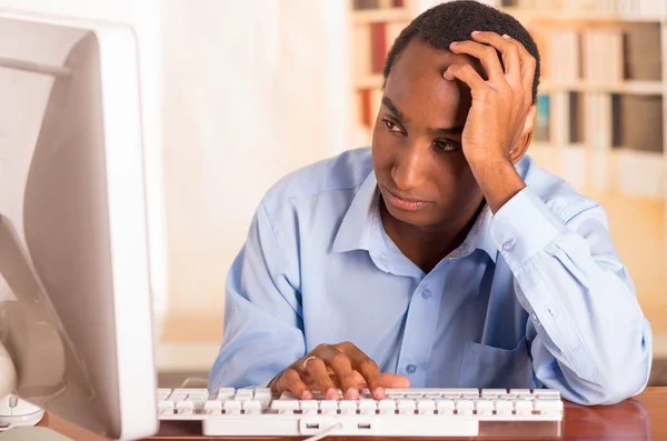 Jovem homem bonito vestindo camisa azul escritório sentado por computador inclinado para a mesa enquanto digita e olhando sem inspiração — Fotografia de Stock