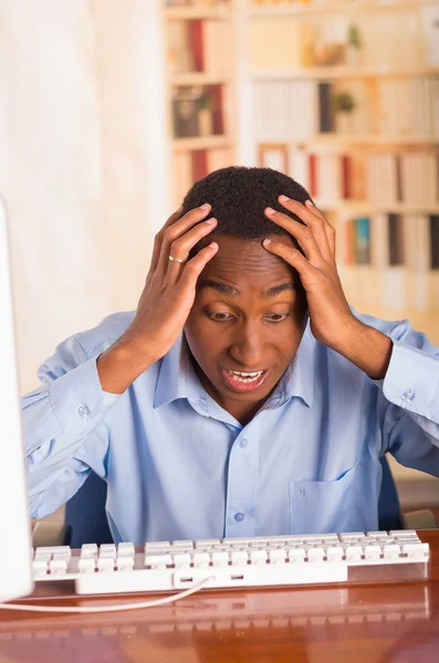 Joven hombre guapo con camisa de oficina azul sentado junto a la computadora inclinado hacia adelante sobre el teclado sosteniendo la cabeza en incredulidad y frustración — Foto de Stock