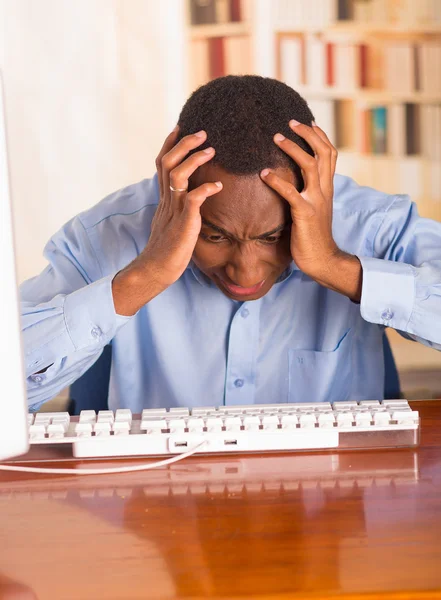 Young handsome man wearing blue office shirt sitting by computer leaning forwards over keyboard holding head in disbelief and frustration — Stock Photo, Image