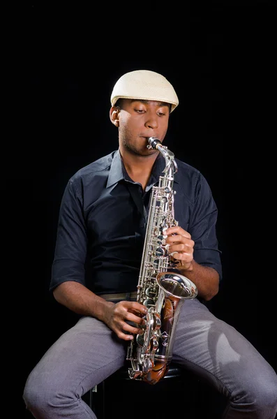 With white hat a black man is sitting and playing his saxophone, dark background, nice music — Stock Photo, Image