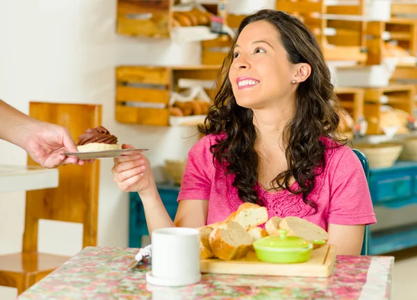 Mulher morena bonita sentada à mesa dentro da padaria, recebendo prato com pão de chocolate e sorrindo — Fotografia de Stock