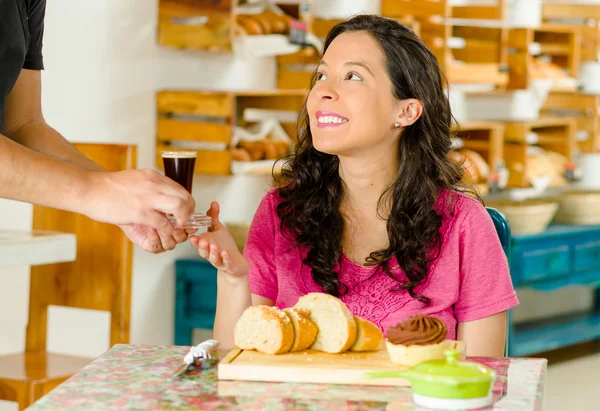 Jolie femme brune assise à côté de la table à l'intérieur de la boulangerie, recevant un expresso du serveur et souriant joyeusement — Photo