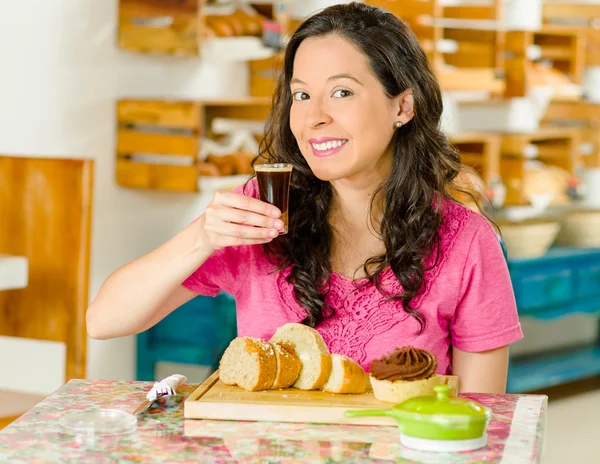 Mujer morena bonita sentada junto a la mesa dentro de la panadería, sosteniendo el vaso de café caliente y posando para la cámara — Foto de Stock