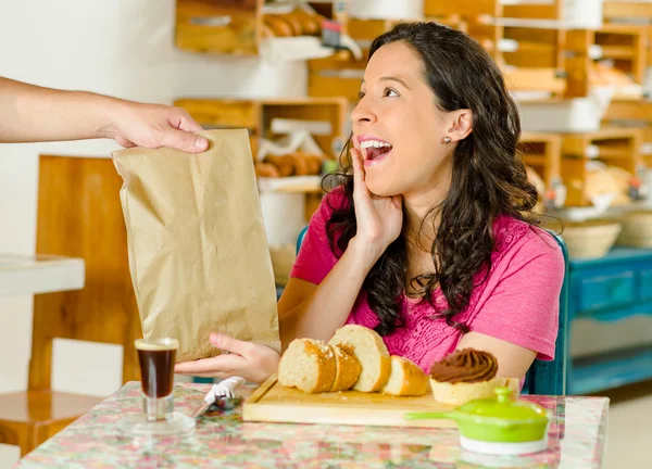 Hübsche brünette Frau in rosa Hemd sitzt am Tisch in der Bäckerei, erhält braune Papiertüte vom Kellner und lächelt glücklich — Stockfoto