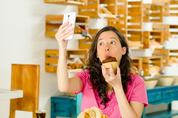 Ładna Brunetka kobieta ubrana w różową koszulę siedząc przy stole wewnątrz piekarni, trzymając telefon komórkowy przy selfie — Zdjęcie stockowe