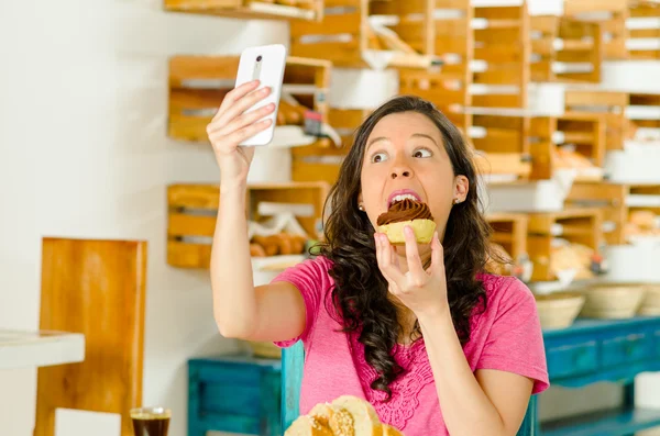 Mujer morena bonita con camisa rosa sentada junto a la mesa dentro de la panadería, sosteniendo el teléfono móvil tomando una selfie —  Fotos de Stock