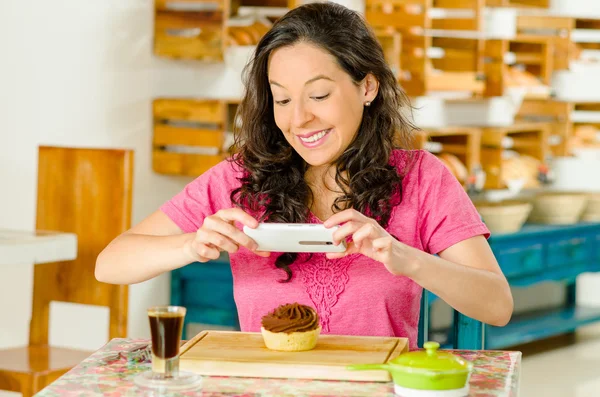 Bella donna bruna che indossa una camicia rosa seduta al tavolo all'interno della panetteria, utilizzando il telefono cellulare per scattare foto di fetta di pane con cioccolato — Foto Stock