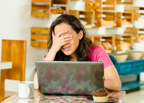 Bella donna bruna che indossa una camicia rosa seduta al tavolo all'interno della panetteria, guardando lo schermo del computer portatile che si copre gli occhi con la mano — Foto Stock