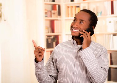 Yukarı doğru işaret ederken cep telefonu konuşurken rahat giysiler giyen yakışıklı adam, beyaz kitaplıklar arka plan