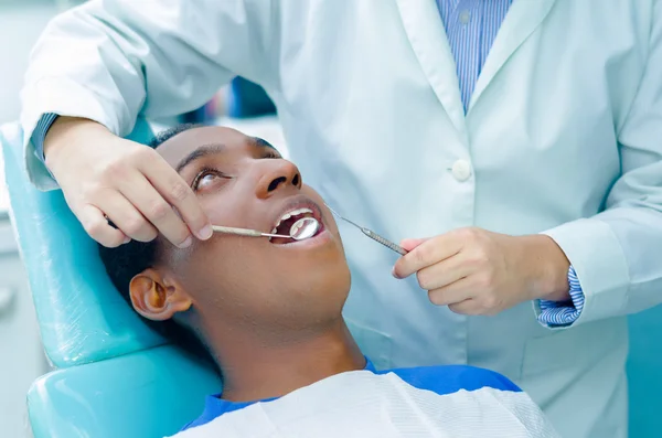 Jeune homme hispanique allongé sur une chaise recevant un traitement dentaire avec la bouche ouverte, les mains de dentiste portant des gants tenant des outils travaillant sur les dents des patients — Photo