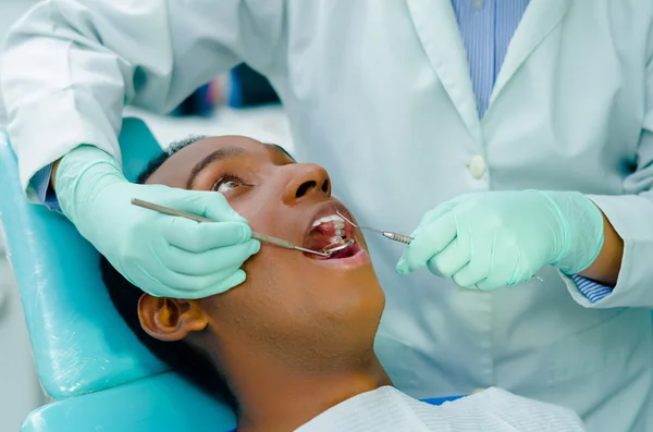 Jonge Latijns-Amerikaanse man in stoel die tandheelkundige behandeling met open mond krijgt, tandartshanden die handschoenen dragen die werktuigen vasthouden die op tanden van patiënten werken — Stockfoto