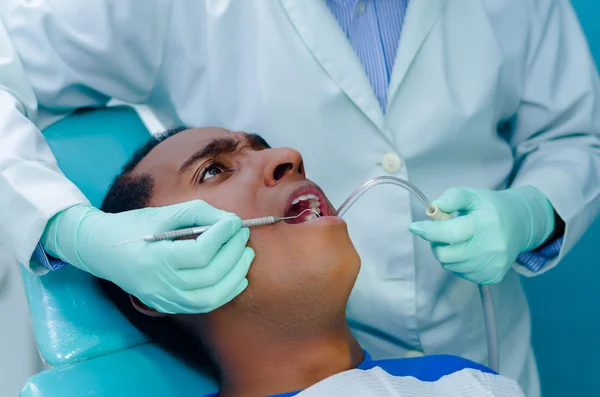 Young hispanic man lying in chair receiving dental treatment with mouth open, dentist hands wearing gloves holding tools working on patients teeth