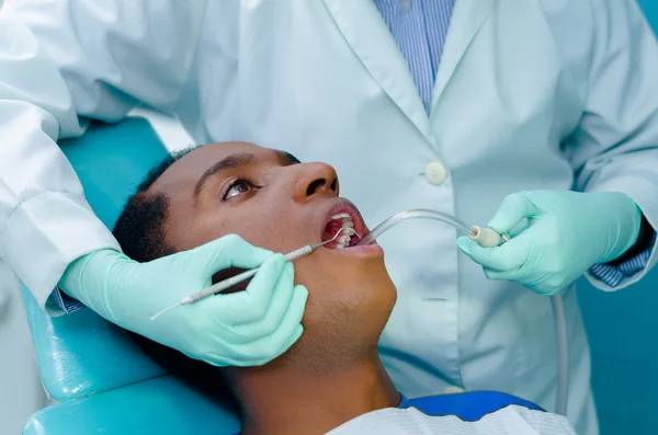 Jeune homme hispanique allongé sur une chaise recevant un traitement dentaire avec la bouche ouverte, les mains de dentiste portant des gants tenant des outils travaillant sur les dents des patients — Photo