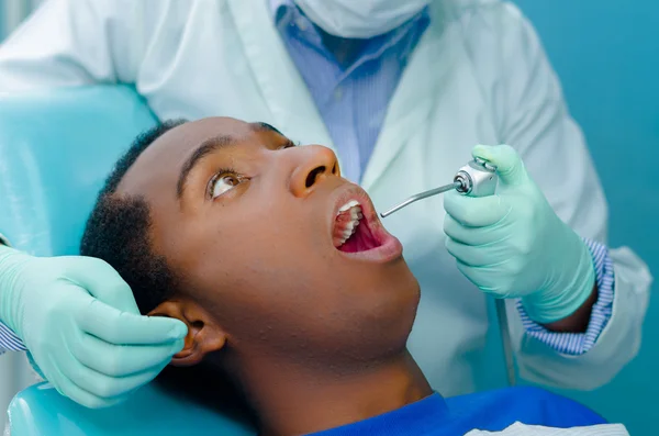 Jeune homme hispanique allongé dans une chaise dentaire levant les yeux, les mains des dentistes avec des gants tenant l'outil dans la bouche des patients — Photo