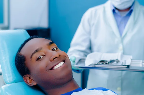 Młody latynoski mężczyzna leżący na krześle dentystycznym patrzący na uśmiechniętą kamerę, płaszcz dentystyczny widoczne tło — Zdjęcie stockowe