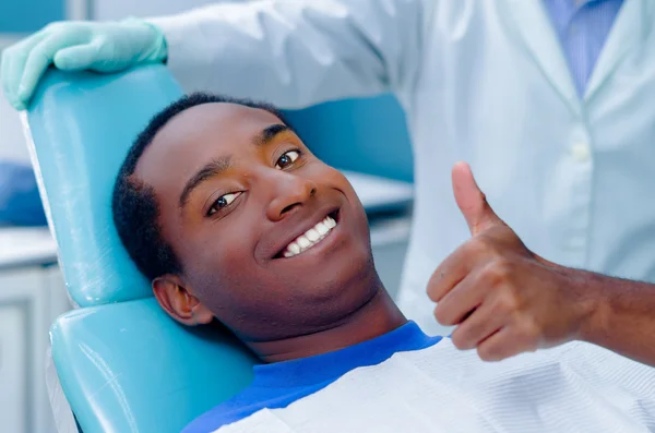 Jeune homme hispanique allongé dans une chaise dentaire regardant la caméra sourire donnant pouce vers le haut, manteau dentiste fond visible — Photo