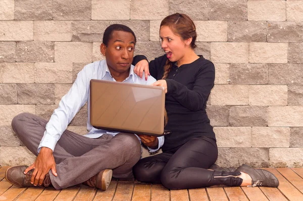 Charmante junge interrassische Paar sitzt an Ziegelwand mit Laptop interagieren und Spaß haben — Stockfoto