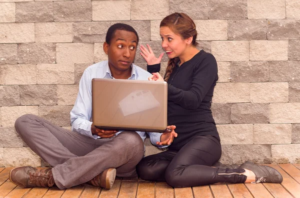 Charming young interracial couple sitting by brick wall with laptop interacting and having fun — Stock Photo, Image