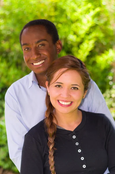 Beautiful young interracial couple in garden environment, embracing and smiling happily to camera — 스톡 사진