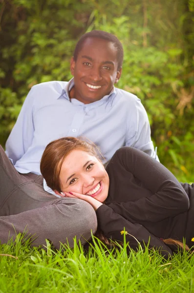 Beau jeune couple interracial dans un environnement de jardin assis, embrassant et souriant joyeusement à la caméra — Photo
