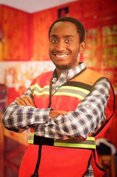 Young carpenter worker wearing square pattern flanel shirt and red safety vest, posing with arms crossed, smiling to camera — Stock Photo, Image
