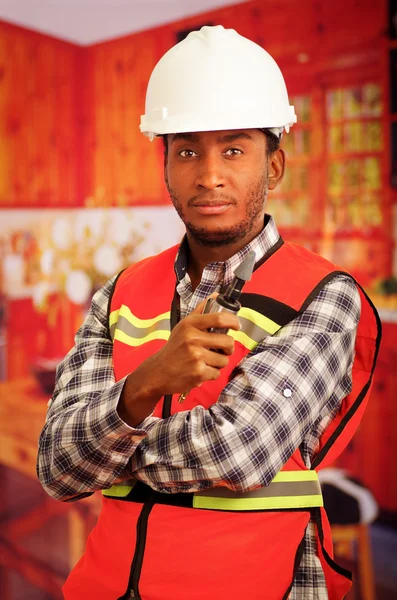 Jovem engenheiro carpinteiro usando capacete, camisa flanel padrão quadrado com colete de segurança vermelho, segurando pequena ferramenta polidor elétrico portátil sorrindo para a câmera — Fotografia de Stock