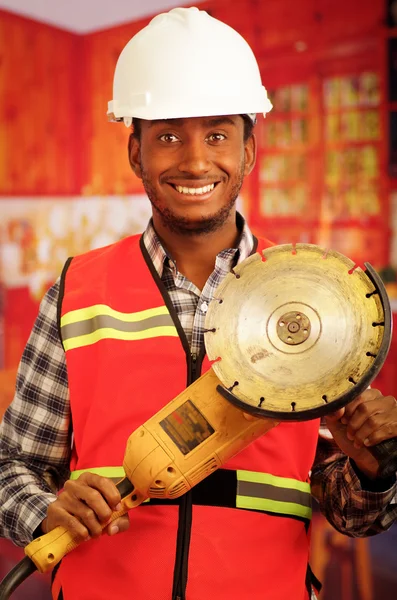 Νεαρός μηχανικός ξυλουργός φορώντας κράνος, τετράγωνο μοτίβο φανέλα πουκάμισο με κόκκινο γιλέκο ασφαλείας, κρατώντας φορητό ηλεκτρικό εργαλείο τριβείο χαμογελώντας στην κάμερα — Φωτογραφία Αρχείου